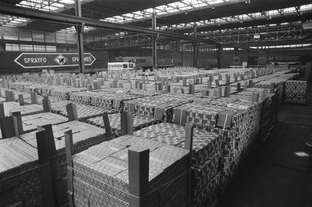 Lagerhalle mit zahlreichen gestapelten Kisten auf dem Boden, metallenen Stützpfählen, Informationsschildern, einem Metalldach mit Beleuchtung und einem geparkten Fahrzeug.