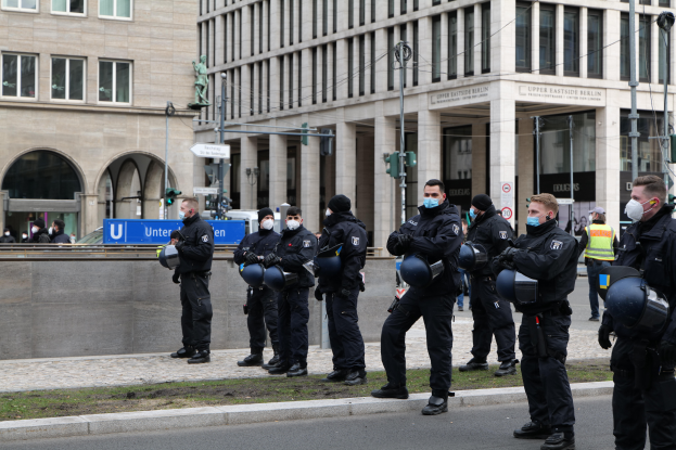 Eine Gruppe von Polizisten in schwarzen Uniformen und Masken steht vor einem Gebäude mit Glasfenstern und Säulen in Berlin, Deutschland, einige halten Helme, mit Laternenmasten, Verkehrsampeln, Texttafeln, einer Statue, anderen Menschen und einem grasbewachsenen Boden im Hintergrund.