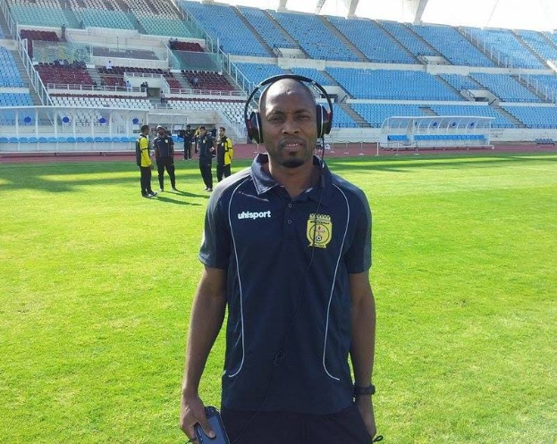 Ein Mann mit Headset, wahrscheinlich der Trainer der ghanaischen Fußballnationalmannschaft, steht auf einem Fußballfeld in der Nähe eines Stadions und hält ein Objekt, während im Hintergrund Menschen zu sehen sind.
