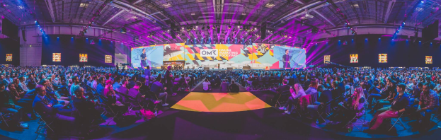 Eine große Menschenmenge sitzt auf Stühlen vor einer hell erleuchteten Bühne mit mehreren Personen, die umgeben sind von Bildschirmen, wahrscheinlich bei einem Online-Marketing-Rockstars-Event.