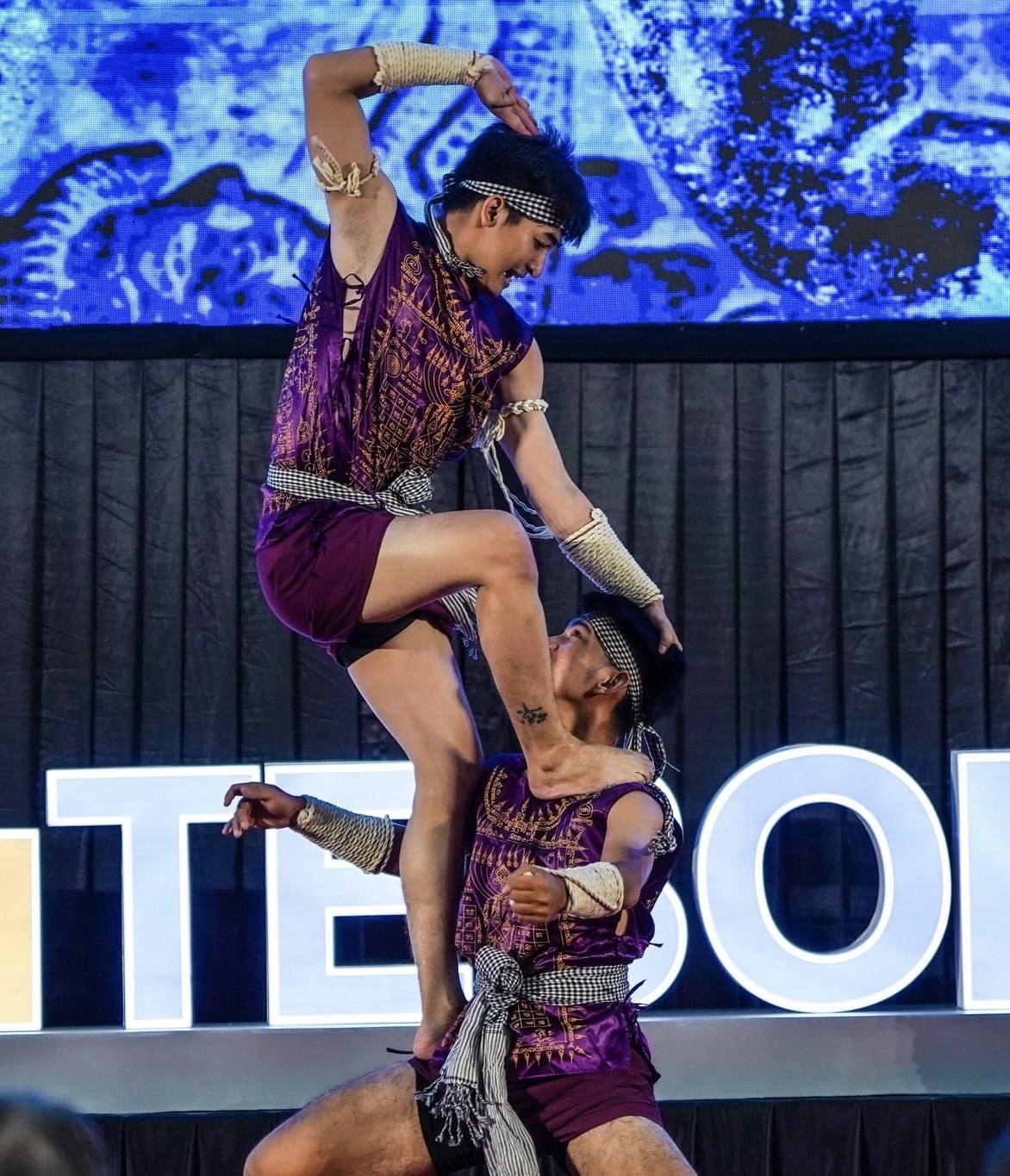 Zwei Akrobaten führen auf der Bühne eine Darbietung auf, wobei der Mann die Frau in lila Kleidern hochhebt, während das Publikum unten zusieht.