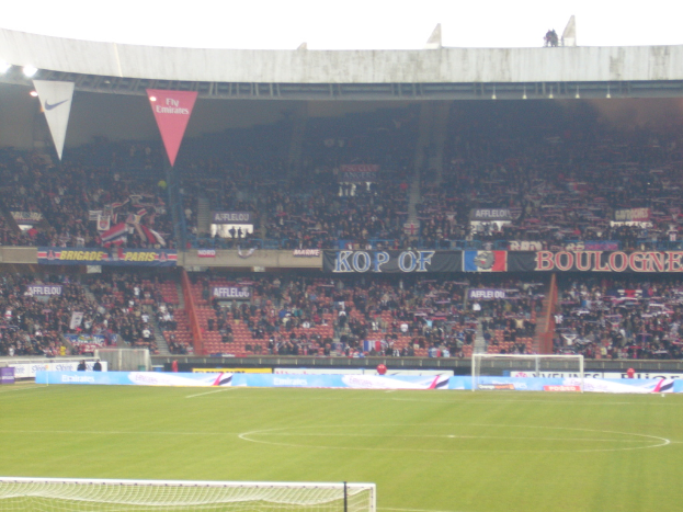 Ein Rasenstadion mit Torpfosten auf beiden Seiten, Zuschauern im Hintergrund und zahlreichen Werbetafeln in der Mitte und darüber.