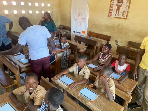 Eine Gruppe von Kindern sitzt an Tischen in einem Klassenzimmer und nutzt Tablets, umgeben von ein paar Menschen, mit einem Banner und Plakat im Hintergrund.