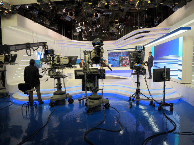 Ein TV-Studio mit mehreren Kameras auf dem Boden, einer sitzenden Person im Vordergrund und einigen stehenden Personen im Hintergrund, ausgestattet mit deckenmontierten Lampen für eine Live-Übertragung.