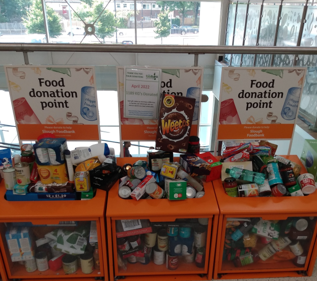 Orange Schübe mit Lebensmittelspenden gefüllt, darunter Flaschen, Dosen und Kisten, mit Informationsschildern in der Nähe, durch Glasfenster mit Außenfahrzeugen, Bäumen, Gebäuden und Himmel.