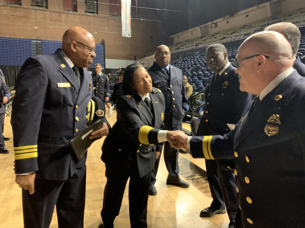 Gruppe uniformierter Personen, die sich die Hände schütteln, vor einem sitzenden und stehenden Publikum, mit einem Banner im Hintergrund, das "Feuerwehrchef und -offizier des Jahres" lautet.