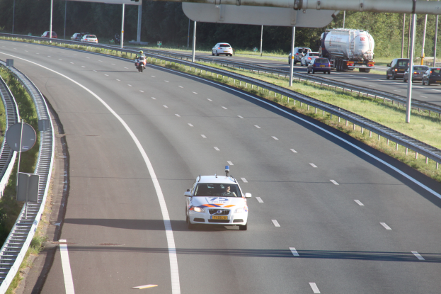 Ein Auto fährt auf einer Autobahn mit einem Polizeiwagen am Röstrand, anderen Fahrzeugen, Leitplanken, Pfosten, Schildern und Bäumen im Hintergrund.