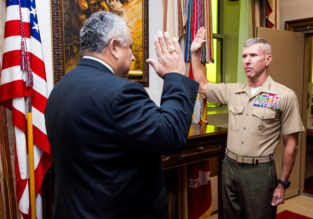 Zwei Männer in Militäruniformen stehen vor Flaggen, mit einem Foto Rahmen an der Wand dahinter; eine Tür und ein Tisch mit verschiedenen Gegenständen sind auf der rechten Seite des Bildes sichtbar.