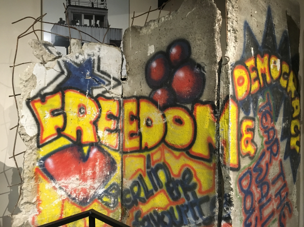 Eine Wand mit mutiger, schwarzer Graffiti-Beschriftung "Freiheit und Demokratie" in einem Raum, mit einem Geländer unten und einem Gebäude mit Fenstern im Hintergrund.