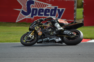 Eine Person in Helm und Schuhwerk fährt ein Motorrad auf einer Rennstrecke, mit Gras und einer Tafel mit Text im Hintergrund.