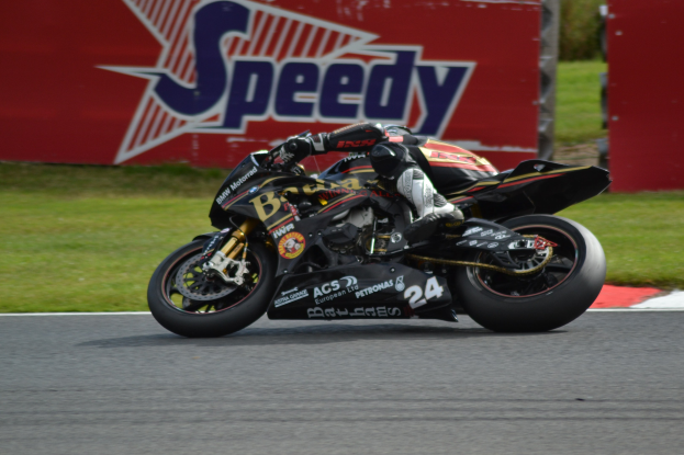 Eine Person in Helm und Schuhwerk fährt ein Motorrad auf einer Rennstrecke, mit Gras und einer Tafel mit Text im Hintergrund.