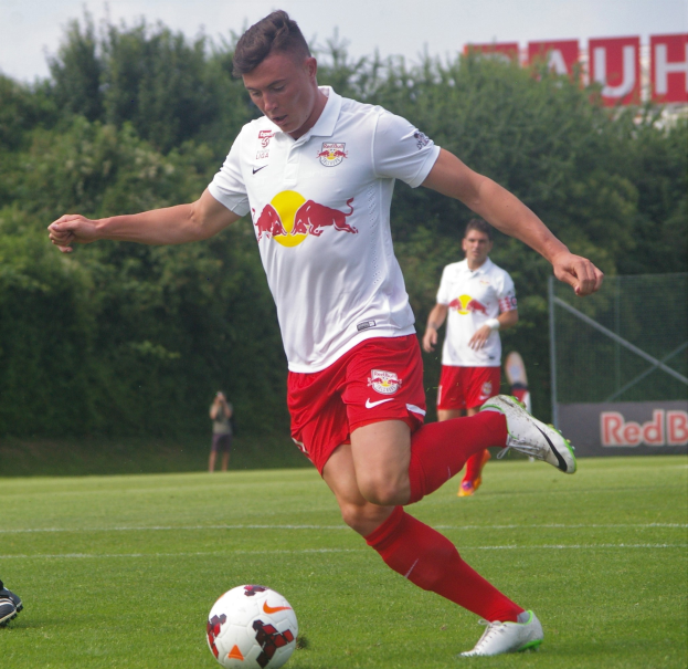 Ein Mann in einem Fußballtrikot kickt einen Ball auf einem grünen Feld mit Bäumen und einem klaren blauen Himmel im Hintergrund, mit einem Schild mit der Aufschrift "RB Leipzig" zu sehen.