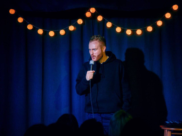 Ein Mann steht auf einer Bühne und hält ein Mikrofon in der Hand, mit einem Tisch voller Gegenstände rechts von ihm und einem sichtbaren Publikum darunter, was auf ein Comedy-Event hindeutet.