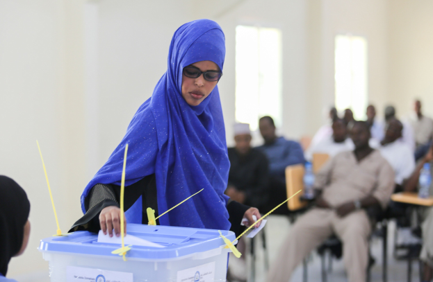 Eine Frau mit einem blauen Kopftuch stimmt in einer Wahllokal ab, während andere in der Nähe sitzen.