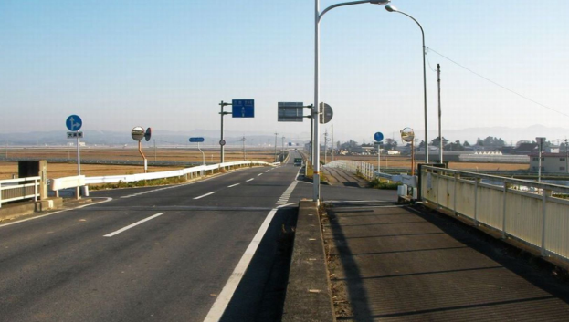 Eine Autobahn mit einer Brücke mit Gelöndern, Laternenmästen und Verkehrszeichen, mit Gebäuden, Bäumen, Höhen und einem klaren blauen Himmel im Hintergrund.