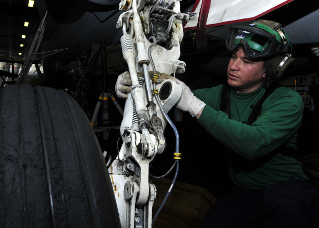 Ein Mann in einem grünen T-Shirt, Helm, Schutzbrille und Handschuhen arbeitet an einem Flugzeugtriebwerk in einer Halle und hält ein Objekt in der Hand, wobei ein Reifen auf der linken Seite zu sehen ist.
