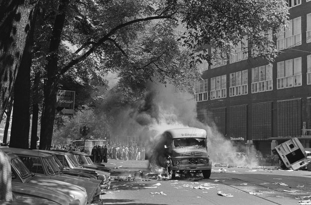 Schwarz-weiß-Foto eines brennenden Busses auf einer Straße mit parkenden Autos auf beiden Seiten, Menschen auf dem Gehweg, Bäume, Gebäude mit Fenstern und einer Reklamewand im Hintergrund.