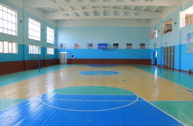 Eine große Turnhalle mit einem blauen und weißen Boden, der einen Basketballplatz, ein Netz mit Pfosten, eine Tür, Fenster, Plakate an den Wänden, ein Geländer und ein Dach zeigt.