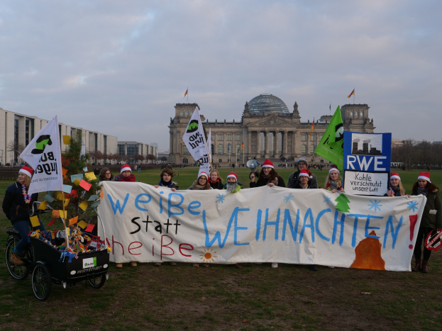 Gruppe von Menschen in Mützen, die ein Banner vor dem Reichstag halten, mit einer Person auf einem geschmückten Kinderwagen, grasbewachsenem Boden, Bäumen, Gebäuden, Flaggen und bewölktem Himmel.