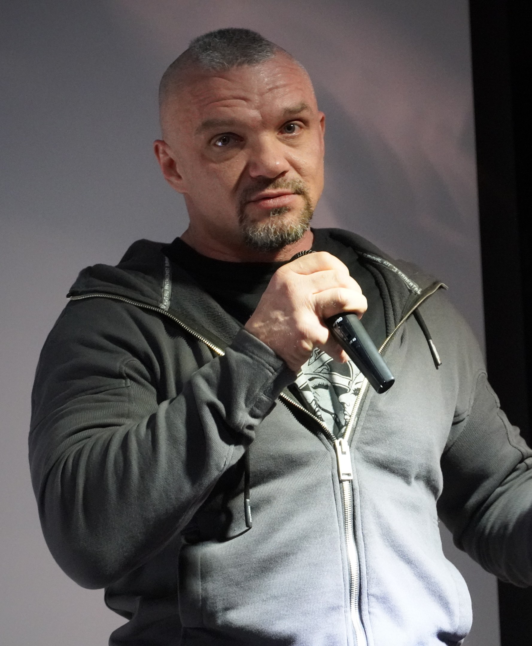 Ein Mann in einer schwarzen Kapuzenjacke und schwarzem Anzug mit einem weißen Hemd, der ein Mikrofon hält und selbstbewusst vor einer Wand steht.