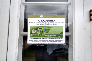 Glasstür mit einem Schild, auf dem "Geschlossene öffentliche Toiletten in der Nähe des Hauptparkplatzes" steht und ein undeutliches Objekt auf der rechten Seite.