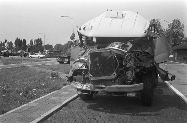 Ein Schwarz-Weiß-Foto eines schwer beschädigten Autos am Straßenrand mit eingedellter Motorhaube und zersplitterten Fenstern, umgeben von anderen Fahrzeugen, Laternenpfählen, Bäumen und einem Haus.