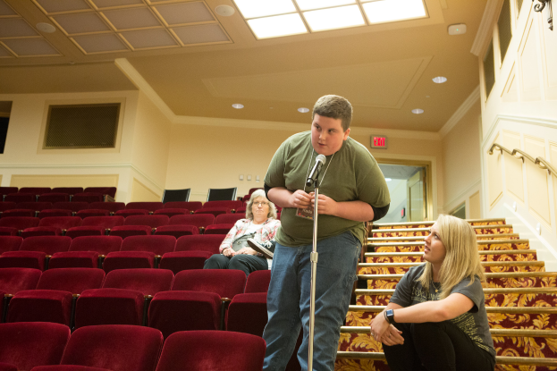 Ein Mann in einem grünen T-Shirt und blauen Jeans steht in einem Theater und hält ein Mikrofon in der Hand, während zwei Frauen auf einer Treppe hinter ihm sitzen, rote Stühle im Hintergrund, ein Geländer auf der rechten Seite und Lampen auf dem Dach.