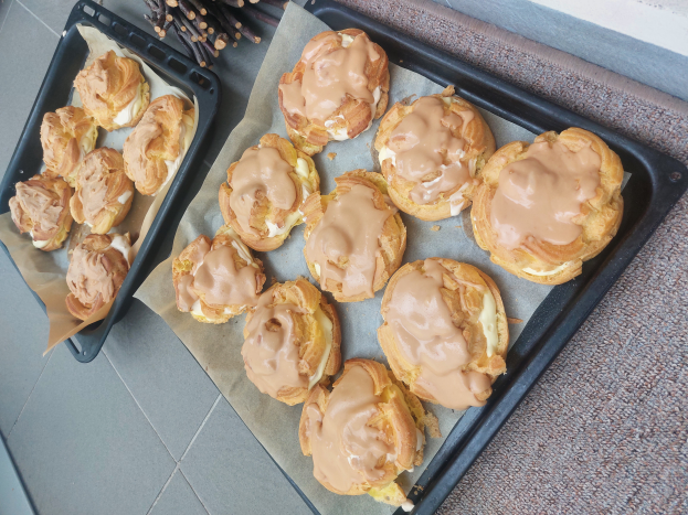 Ein Tablett voller gemischter dänischer Teilchen mit einer hellen goldbraunen Farbe und einer fluffigen Textur, einige mit Puderzucker bestäubt, liegt auf einer Theke neben einem offenen Fenster und wird von warmem Licht beschienen.