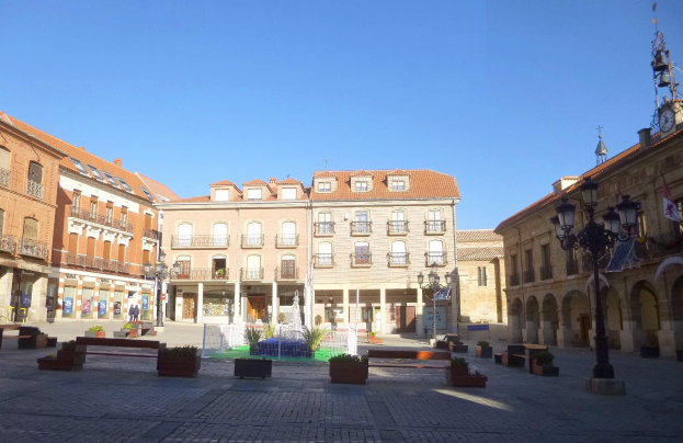 Ein Plaza Mayor in einem Stadtplatz mit einem zentralen Brunnen, umgeben von Bänken, Topfpflanzen, Straßenlaternen, einer Uhrenturm und Gebäuden mit Fenstern, unter einem klaren blauen Himmel.