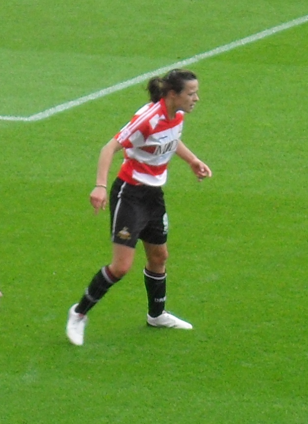 Eine Frau in einem roten und weißen T-Shirt und schwarzen Shorts, die auf einem grünen Fußballfeld spielt und Sportschuhe trägt.