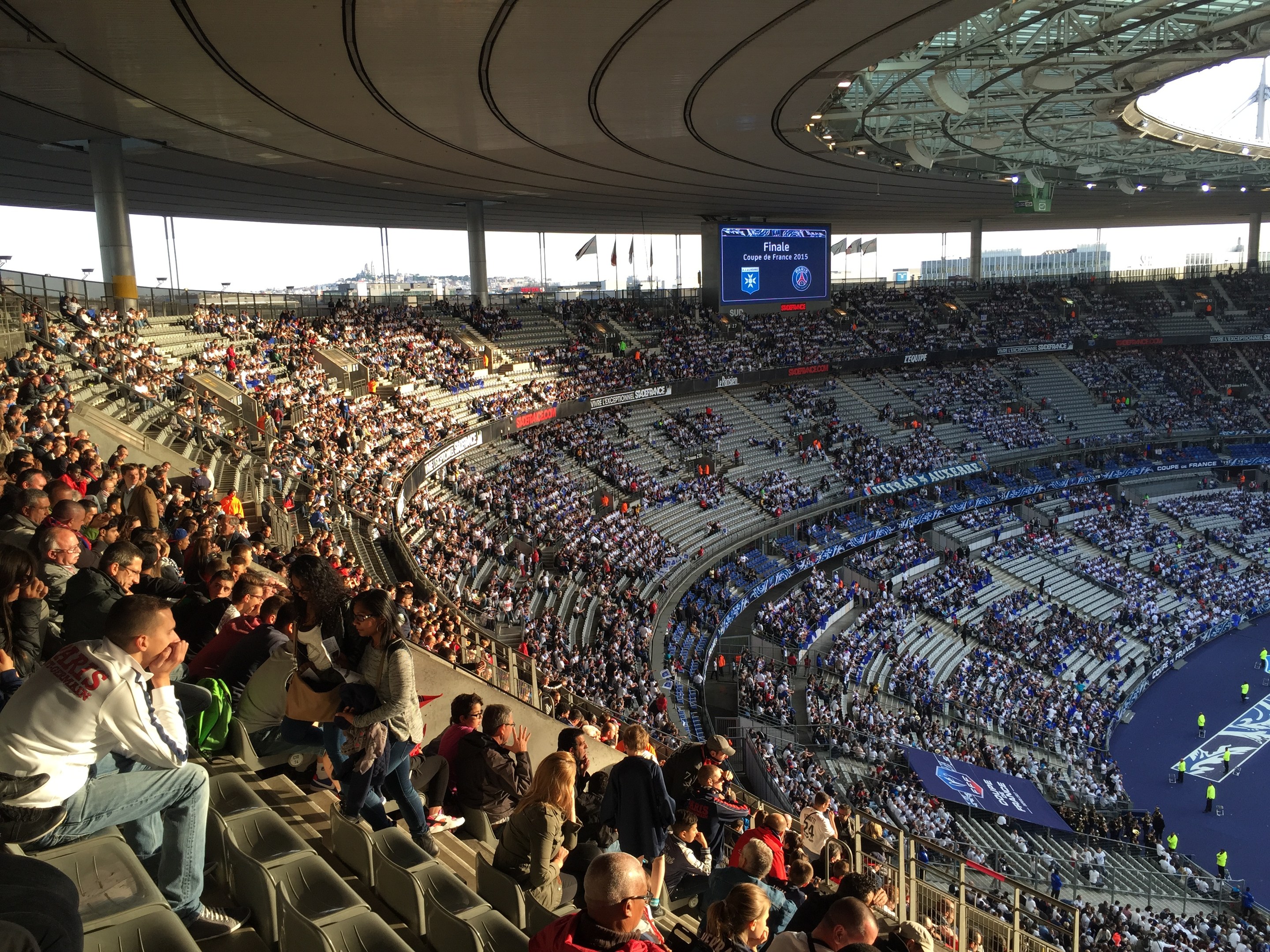 Eine große Menschenmenge in einem Stadion schaut einem Fußballspiel zu, mit einer Bühne, Fahnen, Stangen und einem Bildschirm im Hintergrund.
