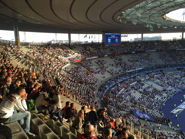 Eine große Menschenmenge in einem Stadion schaut einem Fußballspiel zu, mit einer Bühne, Fahnen, Stangen und einem Bildschirm im Hintergrund.