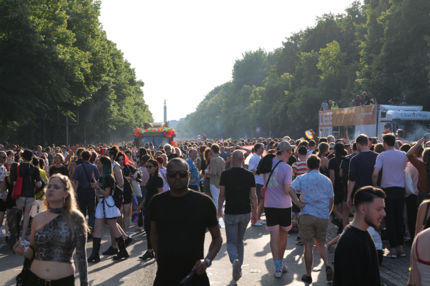 Eine große Menge von Menschen, die eine von Bäumen gesäumte Straße entlanggehen mit einem Turm im Hintergrund, Fahrzeuge mit Menschen auf der rechten Seite, wahrscheinlich beim Christopher Street Day in Berlin unter einem klaren blauen Himmel.