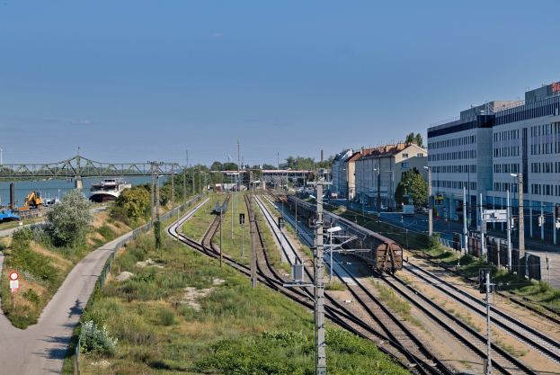 Ein Zug fährt auf Schienen neben einer Stadtlandschaft mit Stromleitungen, Gebäuden, Grünflächen, Verkehrsschildern, Fahrzeugen, einer Brücke, Wasser und einer bewölkten Himmel.