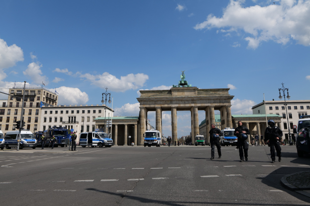 Polizisten vor dem Brandenburger Tor in Berlin, Deutschland, mit Säulen und Statue des Tores, umgeben von Gebäuden, Fahrzeugen, Laternenmasten und Ampeln unter einem bewölkten Himmel.
