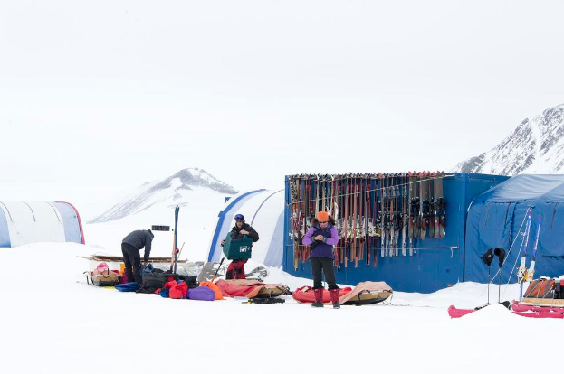 Drei Personen stehen auf einer schneebedeckten Landschaft mit Taschen, die herumliegen, sowie Zelten mit Skiern dahinter und schneebedeckten Hügeln im Hintergrund unter einem klaren Himmel.
