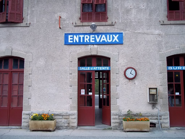 Ein Gebäude mit Türen, Fenstern und einer Uhr an der Wand, mit einem Schild, das Text enthält, Pflanzen und eine Straße unten.