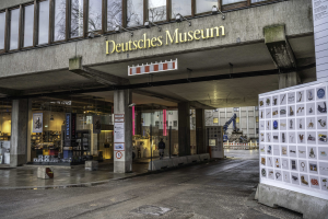 Das Deutsche Museum in Berlin, Deutschland, ein großes Gebäude mit Glasfenstern, Säulen und einer Namenstafel, mit einer Tafel mit Bildern und Text auf der rechten Seite und anderen Gebäuden, Bäumen und dem Himmel im Hintergrund.