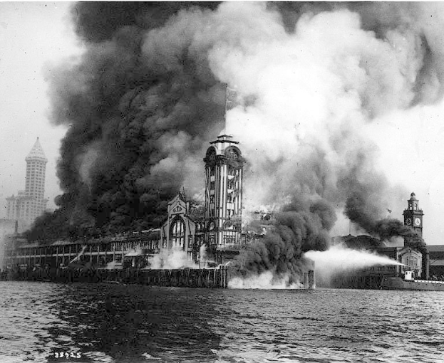 Ein Schwarz-Weiß-Foto eines großen, in Flammen stehenden Gebäudes mit Rauchwolken, einem Gewässer im Vordergrund und mehreren anderen Gebäuden und dem Himmel im Hintergrund.