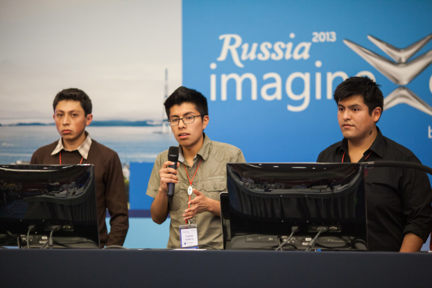Drei Jungen stehen neben einem Schreibtisch mit zwei Computersystemen, einer spricht in ein Mikrofon und trägt ID-Karten mit roten Markierungen, vor einer Wand mit einem Werbeplakat, auf dem "Russia Imagine 2013" steht.