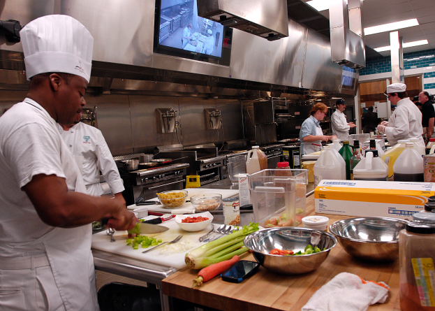 Eine Gruppe von Köchen in einer Küche bei der Zubereitung von Essen auf einem Schneidbrett, wobei ein Koch im Vordergrund in einer weißen Schürze und einer weißen Mütze steht und ein Messer hält und von Utensilien, Schüsseln, Gemüse, einem Handy, Flaschen und anderen Gegenständen umgeben ist, während im Hintergrund ein Fernseher und Deckenlampen zu sehen sind.