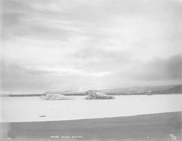 Ein Schwarz-Weiß-Foto eines Eisbergs, der im Ozean treibt, umgeben von Hügeln und einem bewölkten Himmel, mit dem Text 'Sunset Childs Glacier' unten.