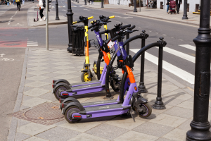 Eine Reihe von E-Scootern, die entlang einer Straße geparkt sind, mit Fußgängern und Radfahrern auf dem Gehweg, einem Müllcontainer im Vordergrund und Gebäuden, Bäumen und Pflanzen im Hintergrund.