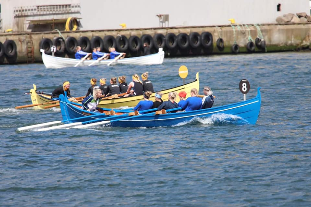 Eine Gruppe von Menschen in einem blauen und gelben Boot auf dem Wasser, die Paddel halten, mit einer Wand mit Reifen und einem Gebäude im Hintergrund, was ein Regatta suggeriert.
