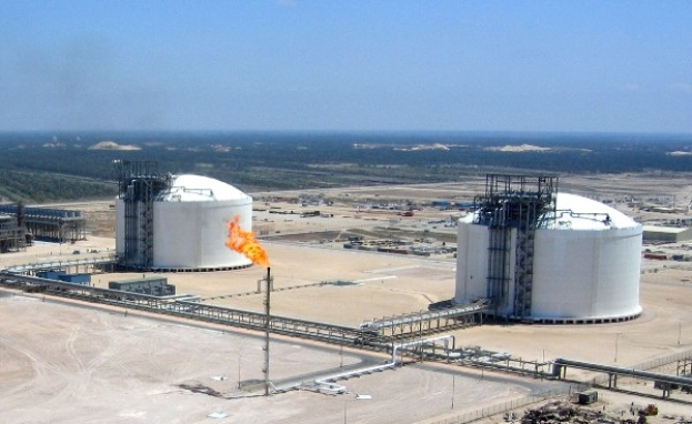 Eine Luftaufnahme eines großeen Ölrefineries mit einem brennenden Feuer in der Mitte, umgeben von zylindrischen Tanks, Polen und anderen Gegenständen, mit Bäumen und einem klaren blauen Himmel im Hintergrund.