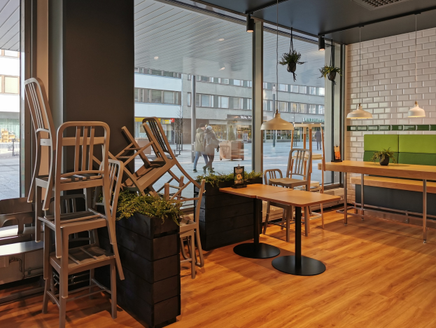 Ein Restaurant in Berlin mit Holztischen und -stühlen vor einem großen Fenster, das urbane Elemente im Freien wie Fußgänger, Gebäude und Straßenmöbel zeigt.