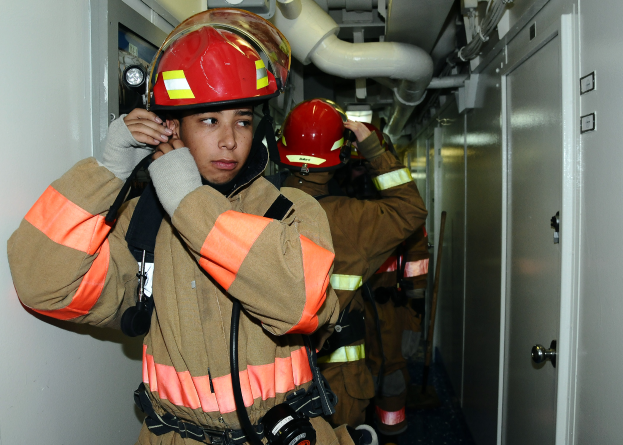 Eine Gruppe von Feuerwehrleuten in Uniform steht zusammen in einem Raum mit einer Tür auf der rechten Seite und einer Wand auf der linken Seite, mit Rohren und anderen Gegenständen im Hintergrund, was auf eine Übung hinweist.
