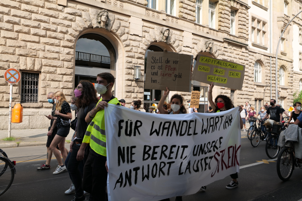 Eine Gruppe von Menschen marschiert auf der Straße in Berlin, hält Schilder und Banner hoch und fährt mit Fahrrädern, vor einem Gebäude mit Fenstern, Bögen, Säulen, Skulpturen, Bäumen und einem Laternenmast, nimmt an einer Anti-Kriegs-Demonstration teil.