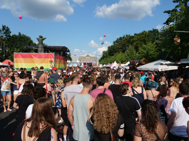 Eine große Menschenmenge, die eine Straße mit Zelten, Bäumen, Pfählen, Lichtern und einer Statue säumt, mit Gebäuden im Hintergrund und einem bewölkten Himmel mit Ballons während des Christopher Street Day in Berlin.