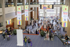 Eine große Menge bewegt sich durch ein großes Gebäude mit Bannern, Topfpflanzen, Rolltreppen, Geländern, Säulen, bedruckten Tafeln, Glastüren und anderen Elementen, einige tragen Taschen bei der Nationalen Buchmesse.